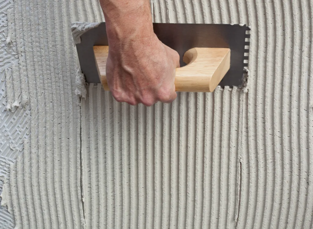 A hand holding a notched trowel applies mortar to a wall surface.