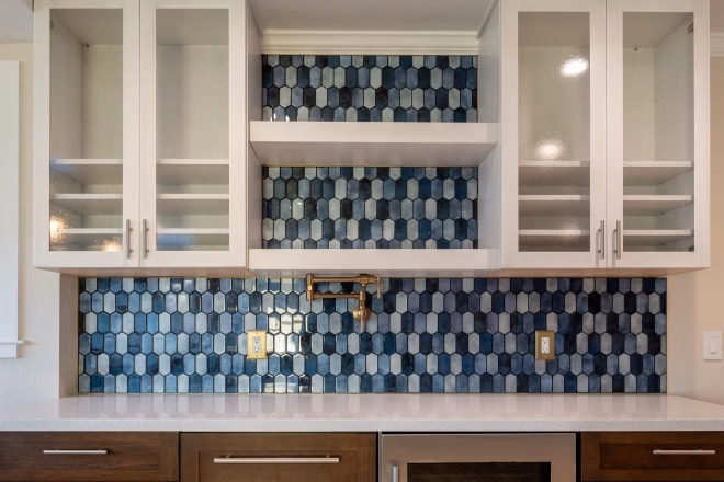 A tile backsplash made of hexagon shaped tiles in various shades of blue with dark wood cabinets underneath and a wine fridge