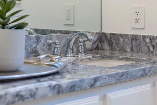 Modern bathroom sink details of shiny chrome faucet with gray stone counter, white cabinets, plant decor and large mirror.