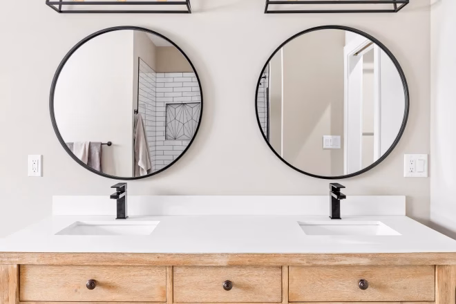 natural wood vanity cabinet, black and gold light fixtures above circular mirrors, and granite countertop